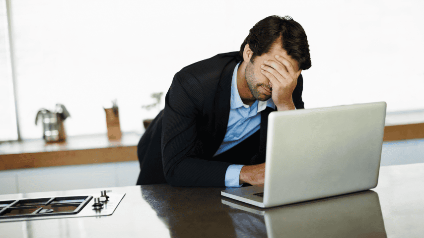 A businessman in a dark suit standing at a kitchen counter with his head in his hand, looking stressed while working on a laptop.